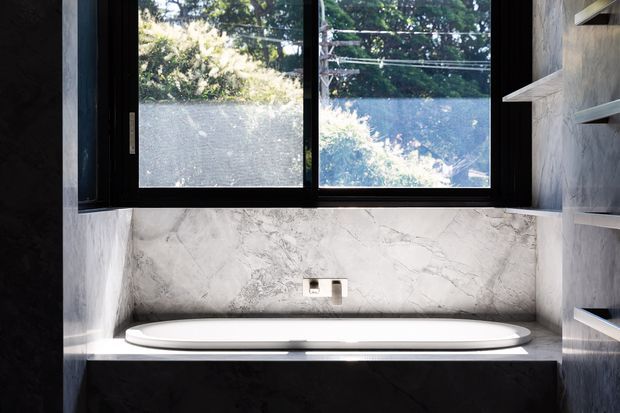 A compact contemporary bathroom featuring a freestanding white bathtub set into a dark stone surround beneath a large black-framed window. Veined light stone extends across the backsplash and side walls, with integrated open shelving on the right and a wall-mounted tap above the tub. Natural greenery is visible outside the window.