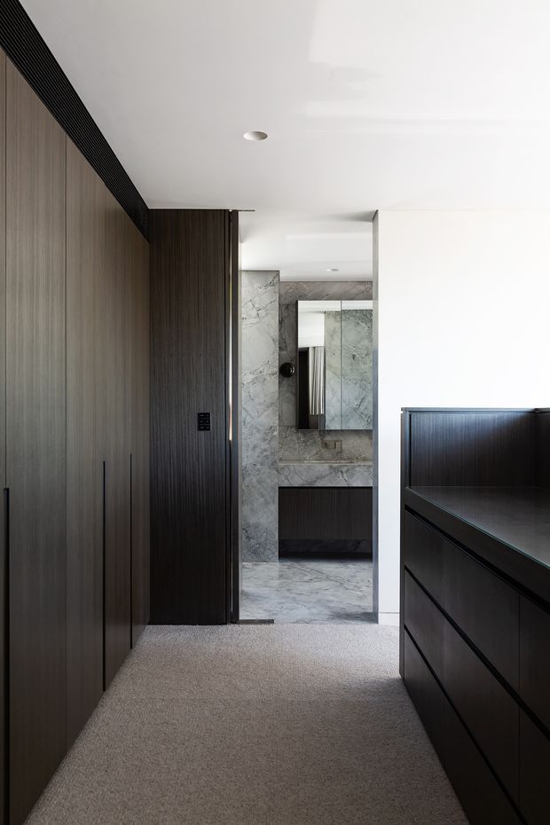 A refined, contemporary dressing area or hallway viewed toward an open bathroom. Full-height dark timber wardrobes line the left wall, with matching dark built-in drawers and a countertop on the right. The carpeted floor leads to a bathroom finished in veined grey stone, featuring a wall-mounted vanity, mirror and dark timber cabinetry. The space has a white ceiling with recessed downlights and a restrained, high-contrast palette.