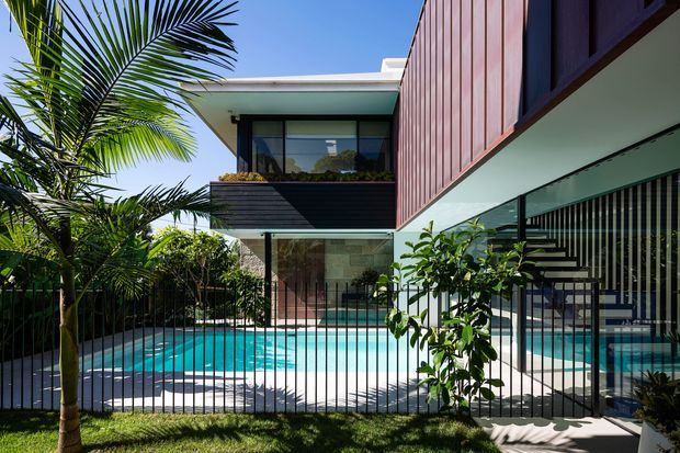 Exterior view of a contemporary multi-level residence centred around a turquoise swimming pool and landscaped courtyard. The building features strong cantilevered volumes, extensive glazing, slim dark metal framing, vertical timber cladding, and a flat roof. A black vertical-bar fence separates the lawn from the pool, while tropical palm planting reinforces the warm outdoor setting.
