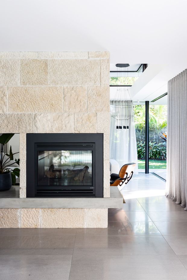 Bright contemporary interior centered on a substantial cream textured stone fireplace with a black built-in firebox. The space has a polished light-grey tiled floor, white ceiling, full-height glazing, sheer curtains, a sculptural pendant light, and a mid-century-style lounge chair opening toward a lush garden. The open-plan room feels airy and refined, with strong indoor–outdoor connection.