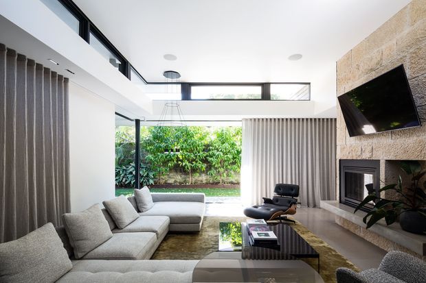 Bright contemporary living room with a large sectional sofa arranged around a low coffee table, a sculptural lounge chair, and a stone-clad fireplace wall with a mounted television. Floor-to-ceiling glazing and high clerestory windows open to a leafy courtyard, while full-height curtains and recessed lighting create a refined, airy interior.