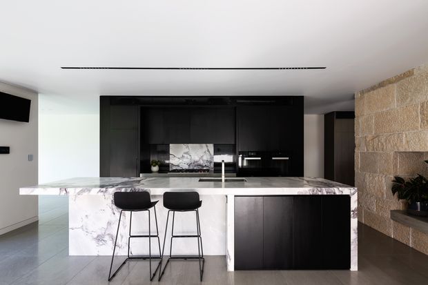 A contemporary kitchen with a large marble-look island and two black bar stools. Dark timber cabinetry forms a full-height kitchen wall, while a substantial exposed stone feature wall with integrated shelving adds texture. The space has a white ceiling, pale floor, integrated appliances, and a minimalist linear ceiling light.