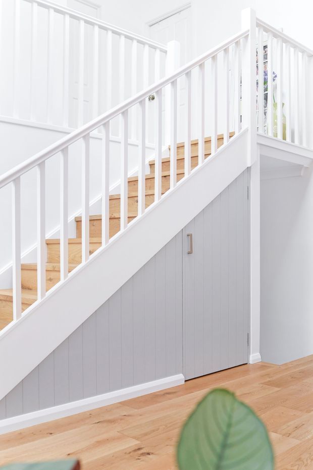 Bright, clean interior view of a white-painted staircase with slender vertical balusters, timber treads, and a grey-white paneled storage cupboard beneath the stairs. Light timber flooring and white wall panelling create a calm, refined aesthetic.
