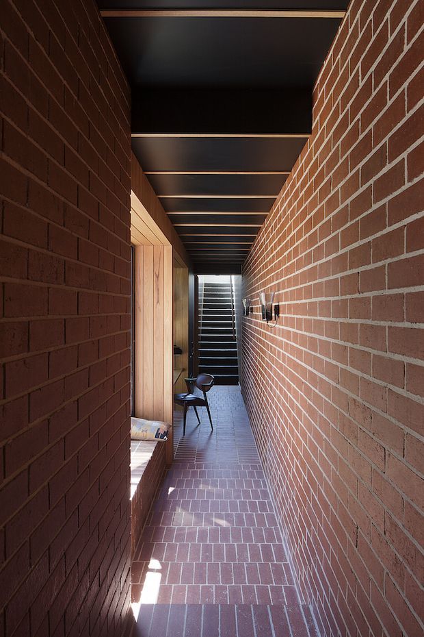 A narrow interior passageway enclosed by exposed red brick walls, with a matching brick-paver floor and a dark ceiling framed by regularly spaced timber beams. A timber-lined opening and built-in seating are visible on the left, with a small chair farther down the corridor and daylight entering at the end. The space has a warm, tactile, contemporary architectural character with strong linear perspective.