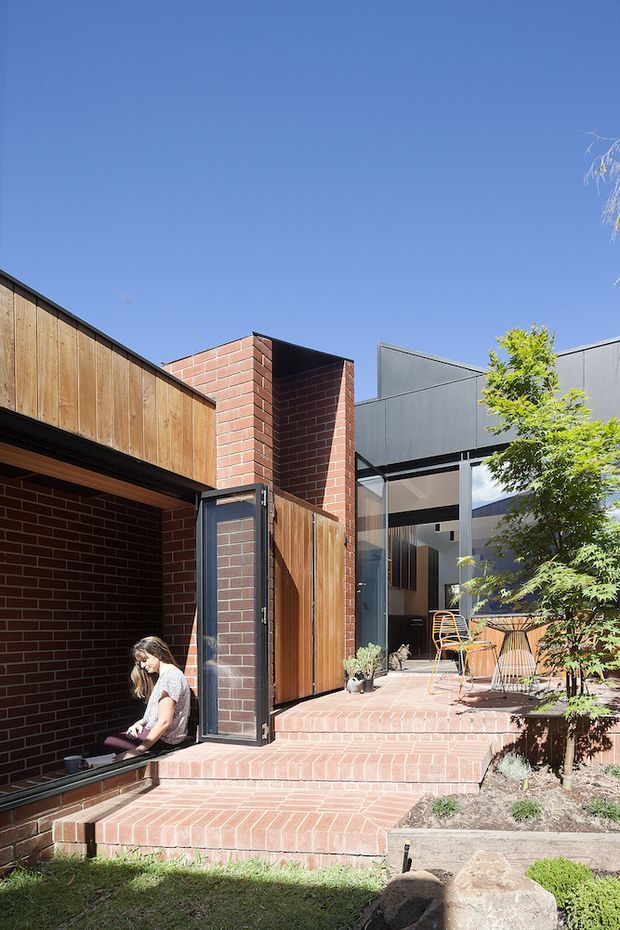 A bright exterior courtyard and entry area of a contemporary brick-and-timber residence. Red brick walls and stepped paving form a recessed entrance, with warm vertical timber cladding, dark metal-framed glazing, and a small landscaped courtyard with a tree and outdoor chairs. Strong geometric forms and clear blue sky create a crisp architectural presentation.