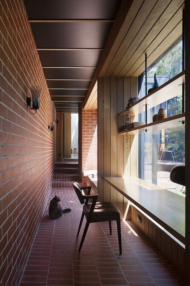 A narrow, warm-toned interior corridor or passage with exposed red brick walls and brick tile flooring. One side features full-height glazing with built-in timber shelving and a long timber ledge or desk, while the ceiling has dark recessed panels with exposed timber beams. Two dark wooden lounge chairs sit along the window, with natural light entering from the garden outside. The space has a strong linear composition and mid-century architectural character.