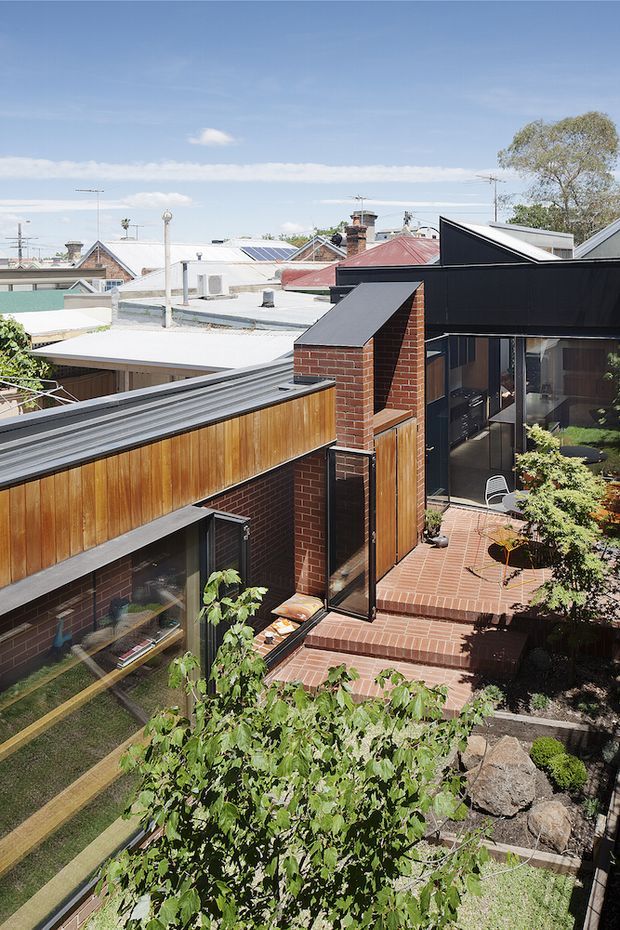 Elevated view of a compact modern residential courtyard and rooftop terrace, featuring dark metal roof forms, exposed red brick walls, warm timber screening, extensive glazing, and terracotta brick paving. The home is integrated into a dense suburban setting with neighbouring rooftops visible beyond.