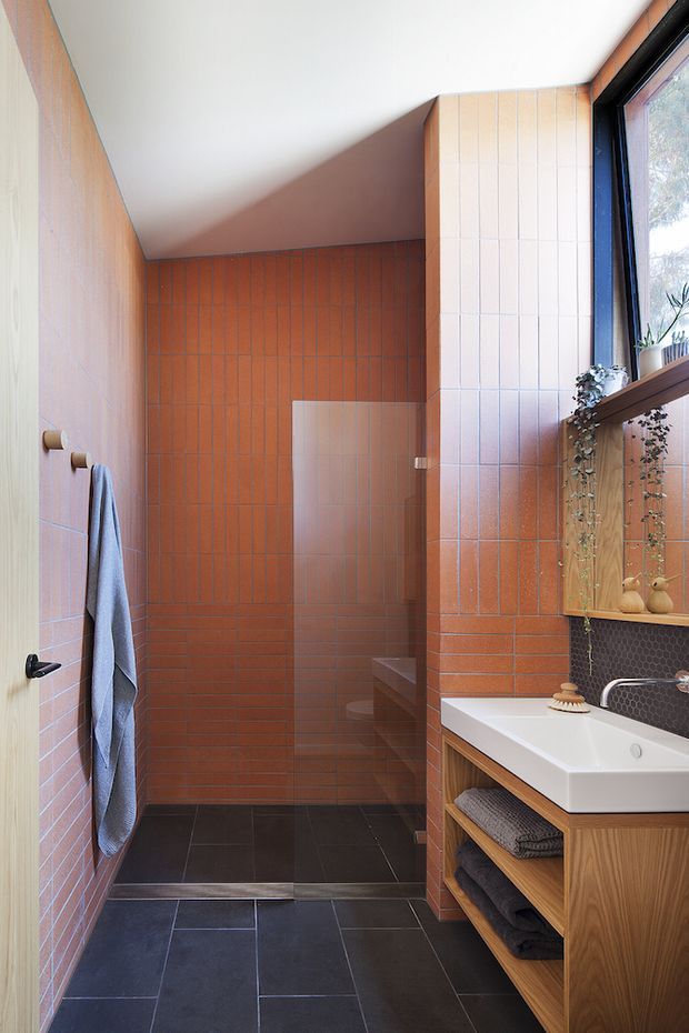 A compact contemporary bathroom with a walk-in tiled shower, wall-mounted towel hooks, a white vessel basin on a timber vanity, open towel storage, and a large window. Warm terracotta vertical wall tiles contrast with dark charcoal floor tiles, white wall tiles, timber cabinetry, and a dark patterned splashback.