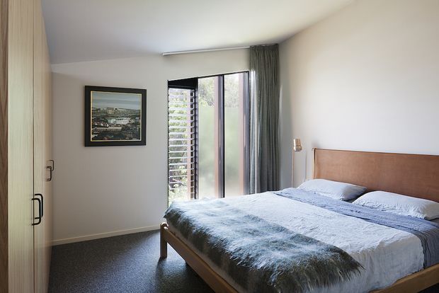 A bright, compact modern bedroom with a timber bed and upholstered tan headboard, layered blue-grey bedding, dark carpet, built-in timber wardrobe storage, and full-height glazed doors with vertical curtains and external louvres. A framed artwork and slim bedside light add restrained decoration.