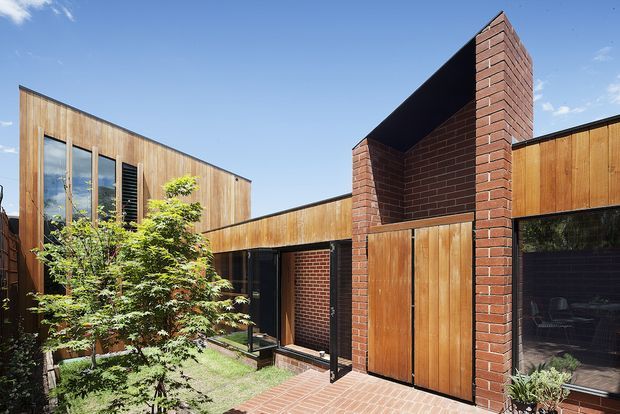 Exterior view of a contemporary multi-volume residence arranged around a small landscaped courtyard. The building combines warm vertical timber cladding, dark-framed glazing, exposed red brick walls, and a tall brick entrance element with timber doors. Brick paving and a Japanese maple contribute to the sheltered courtyard setting, under a clear blue sky.