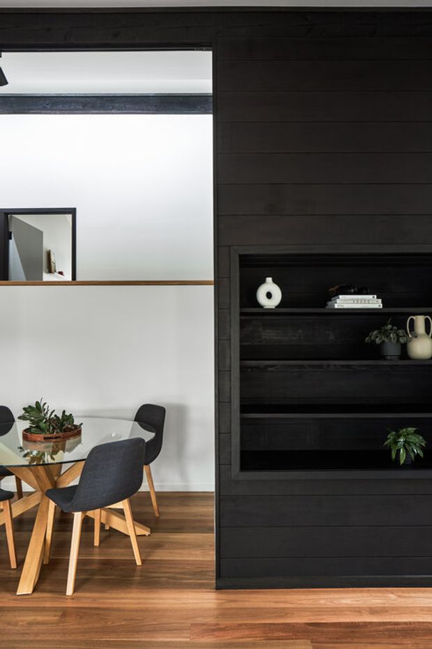 Interior view showing a dining area beside a dramatic dark timber-lined built-in shelving wall. The dining space has a round glass table with dark upholstered timber-leg chairs, a potted plant centerpiece, white walls, and warm timber flooring. A large internal opening with a timber ledge connects visually to an adjacent bright space. The built-in shelving contains minimal decorative ceramics, books, and plants. The composition is contemporary, high-contrast, and warm.