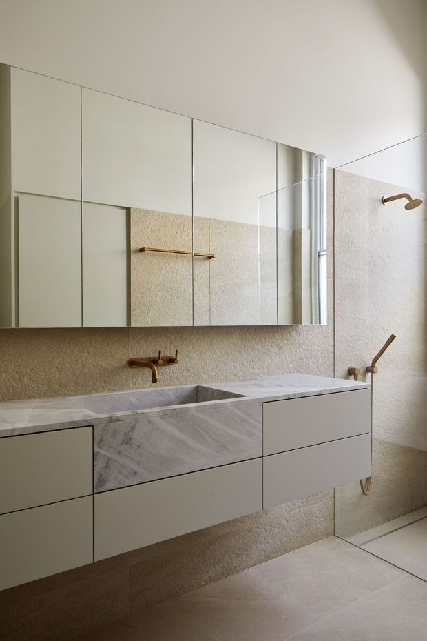 A refined contemporary bathroom featuring a wall-mounted double vanity with pale stone surfaces, integrated basins, and soft off-white drawers. Large mirrored cabinetry spans the wall above a textured beige plaster backsplash, reflecting a brass towel rail and window. Brushed brass wall-mounted taps, a shower fitting, and a curved exposed waste detail add warmth. Large neutral floor tiles and abundant natural light create a calm, luxurious atmosphere.