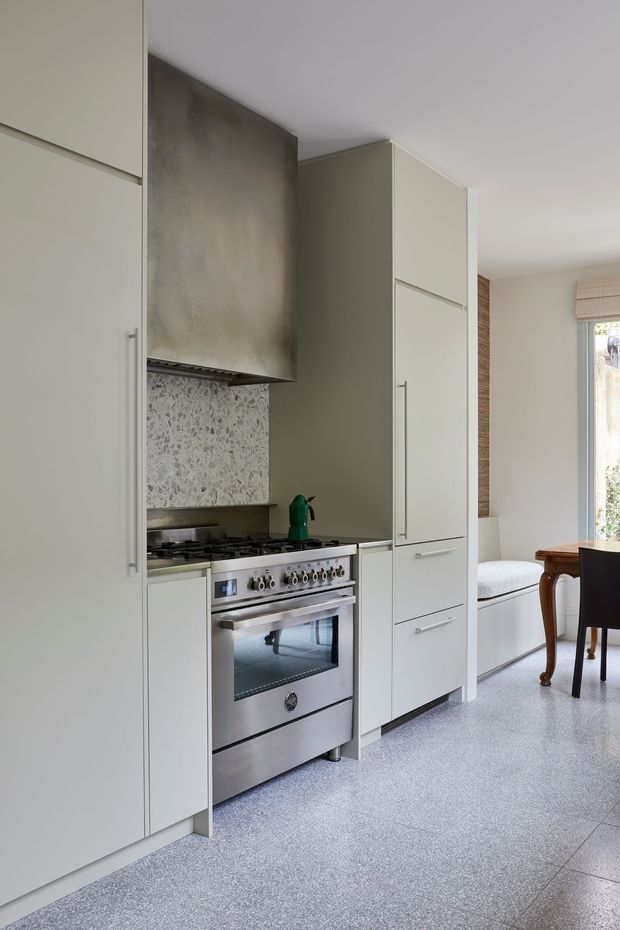 A compact, refined kitchen with full-height pale cabinetry, integrated refrigerator storage, a freestanding stainless-steel range, metallic rangehood, and a light terrazzo-style backsplash. The kitchen opens toward a bright dining area with a timber table, upholstered bench seating, and a window with a woven blind. The palette is soft grey, off-white, and stainless steel, creating a clean contemporary atmosphere.