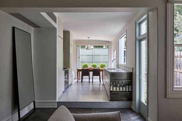 A bright, narrow open-plan kitchen viewed from an adjoining living area. The kitchen features a long stone island with an undermount sink and integrated storage, white cabinetry, tiled flooring, and large windows and glazed doors providing abundant natural light. A dining table sits at the far end, with a small adjacent living area in the foreground. The space has clean contemporary detailing and a compact, elongated layout.