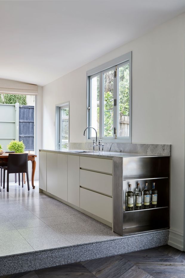 Bright contemporary kitchen with pale built-in cabinetry, a grey marble-look benchtop and splashback, stainless-steel sink and open bottle-storage shelving. The kitchen sits on a raised tiled platform beside a dining area, with large grey-framed windows bringing in abundant natural light. Dark herringbone timber flooring is visible in the foreground.