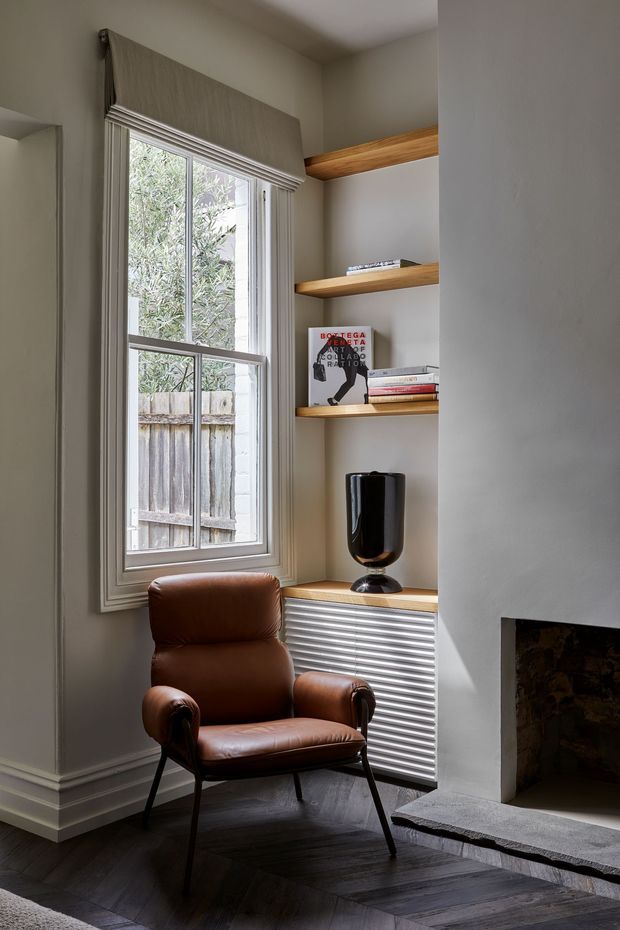 A refined living room corner with a cognac leather lounge chair beside a tall window, built-in timber display shelves, and a concealed radiator cabinet with ribbed metal detailing. A simple black vase, books, and artwork sit on the shelves. The room includes a traditional-style fireplace with a grey stone hearth and dark herringbone timber flooring, combining classic architectural detailing with contemporary furnishings.
