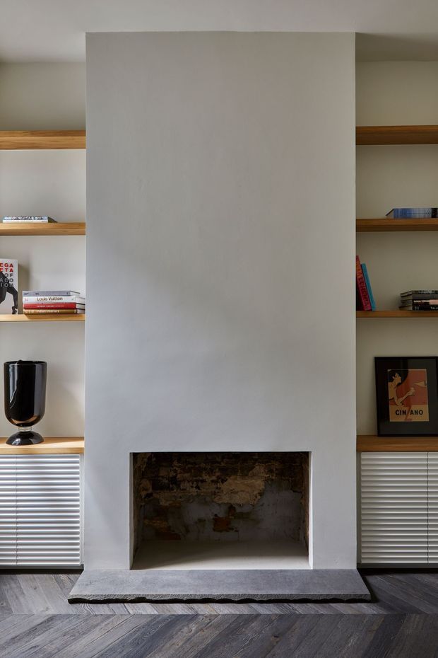 Interior view of a minimalist living room feature wall with a large white plaster fireplace surround and exposed rustic masonry inside the firebox. Open timber display shelves flank the fireplace, holding books and decorative objects, with grey horizontal blinds below. The room has dark grey timber flooring laid in a diagonal or herringbone-like pattern and a restrained, contemporary palette.