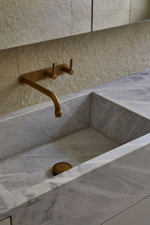 Close-up of a minimalist bathroom vanity featuring a large integrated rectangular stone basin and matching stone countertop and backsplash. The basin and surfaces have a grey veined natural-stone appearance, paired with a wall-mounted brushed bronze/gold three-piece tap set. A frameless mirror and textured stone wall tiles complete the refined, tonal composition.