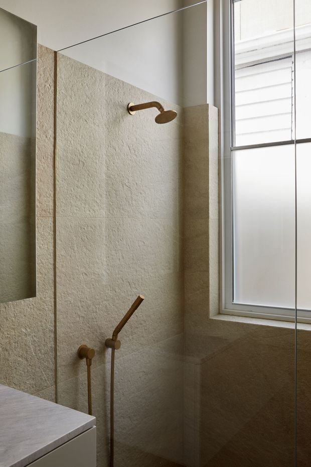 A close-up view of a contemporary walk-in shower within a bathroom. The shower walls are clad in large-format textured beige stone-look tiles, with brushed brass shower fittings and a frameless glass screen. A pale stone vanity edge is visible at the lower left, while a frosted window with white framing provides natural light.