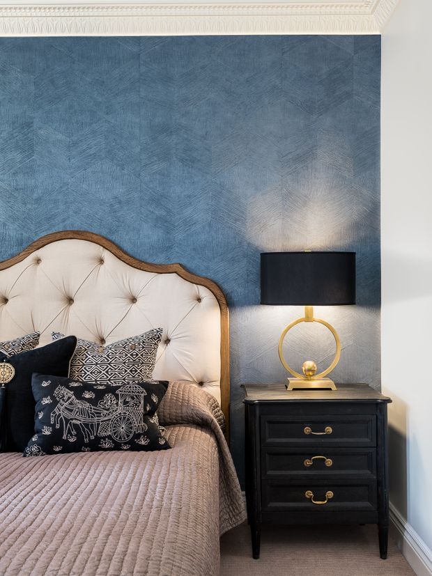 A close-up view of a traditionally styled bedroom featuring an ornate upholstered bedhead with dark timber edging, layered patterned and dark cushions, a quilted taupe bedcover, and a black bedside chest with brass handles. The room has a textured blue feature wall, white decorative cornice moulding, and a circular gold table lamp with a black shade.