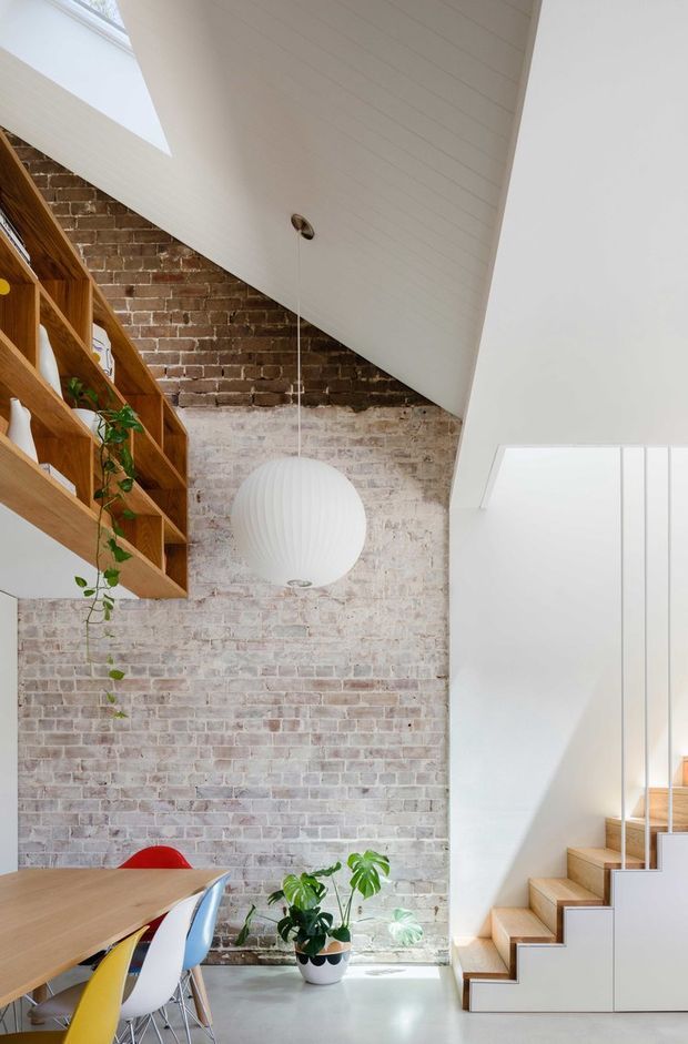 Bright, double-height contemporary interior featuring an exposed pale brick feature wall, timber open shelving, a dining table with colourful chairs, indoor plants, a large white spherical pendant light, and a compact timber staircase with white balustrade and skylight glazing. The space has a loft-like, open-plan character with strong natural light.