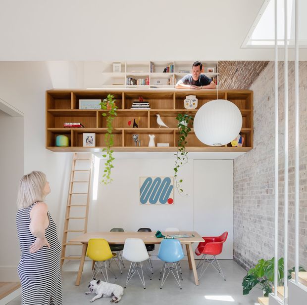 Bright, double-height contemporary dining and living space with a large built-in timber shelving unit spanning the upper wall. A dining table with colourful mid-century-style chairs sits below, alongside a leaning ladder, hanging plants, artwork, and a large spherical pendant light. Exposed white-painted brick forms the right wall, while glazing and a high clerestory/skylight bring in abundant daylight. A person and dog are present but are not central to the architectural composition.