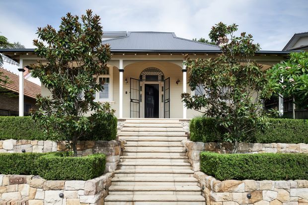 Front exterior view of a refined single-storey residence with a symmetrical covered entry porch, pale rendered walls, dark metal security and entry doors, and a low-pitched corrugated metal roof. A broad flight of pale stone steps rises through formal clipped hedges and layered sandstone retaining walls, with established ornamental trees framing the facade.