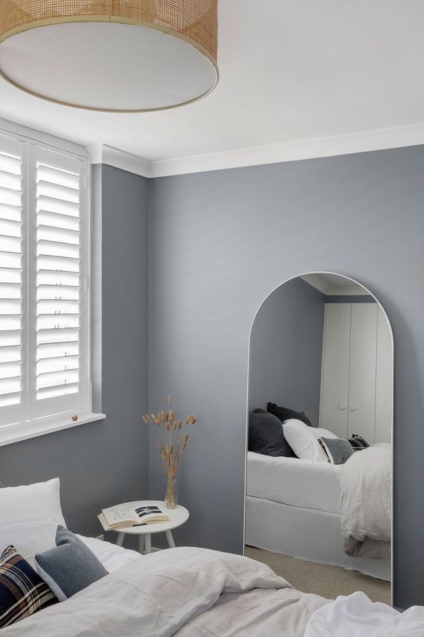 A calm, contemporary bedroom with soft grey walls, white plantation shutters, light carpet, and a partially visible bed dressed in white bedding and patterned cushions. A large arched full-length mirror leans against the wall, reflecting the bed and built-in wardrobe. A woven rattan drum pendant and a small round bedside table with dried branches add warm natural accents.