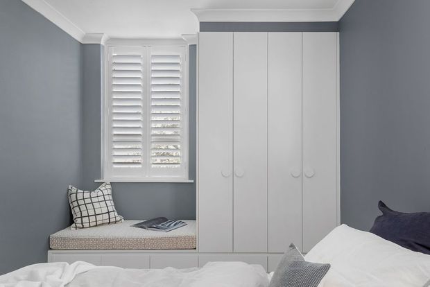 A compact, calm bedroom with dark grey-blue walls, white decorative ceiling trim, a built-in white wardrobe, and a window seat beneath plantation shutters. The bed occupies the foreground with white bedding and neutral patterned cushions, creating a clean, tailored interior.