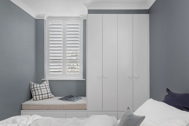A compact, calm bedroom with grey walls, white plantation shutters, built-in white wardrobes, and a window seat with a patterned cushion. The foreground shows part of a bed with white bedding and neutral accent pillows. Clean built-in joinery and restrained tones create a contemporary, refined look.