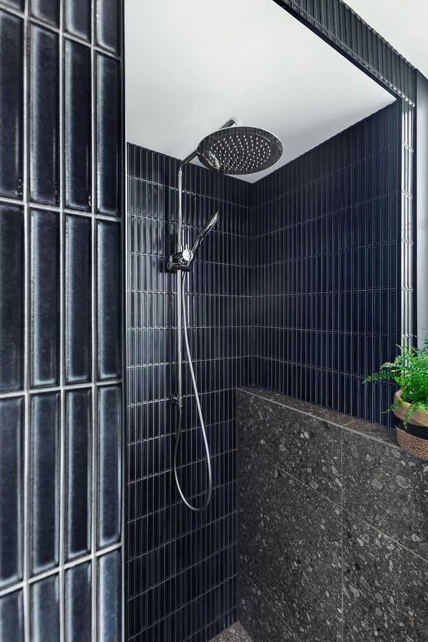 A compact contemporary walk-in shower with glossy dark blue-black narrow-format wall tiles, a large round overhead rain shower, handheld shower fitting, and a raised dark terrazzo-clad partition or bench. A potted plant adds a small natural accent.