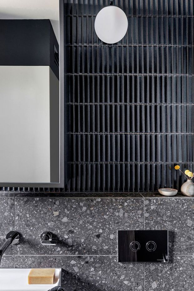 Close-up view of a sophisticated contemporary bathroom vanity wall. The composition features glossy black vertically ribbed tiles, a large frameless mirror with a dark projecting surround, dark terrazzo wall cladding, black wall-mounted tapware, and a black dual-flush plate. Small neutral decorative vessels and a white soap dish add subtle contrast.