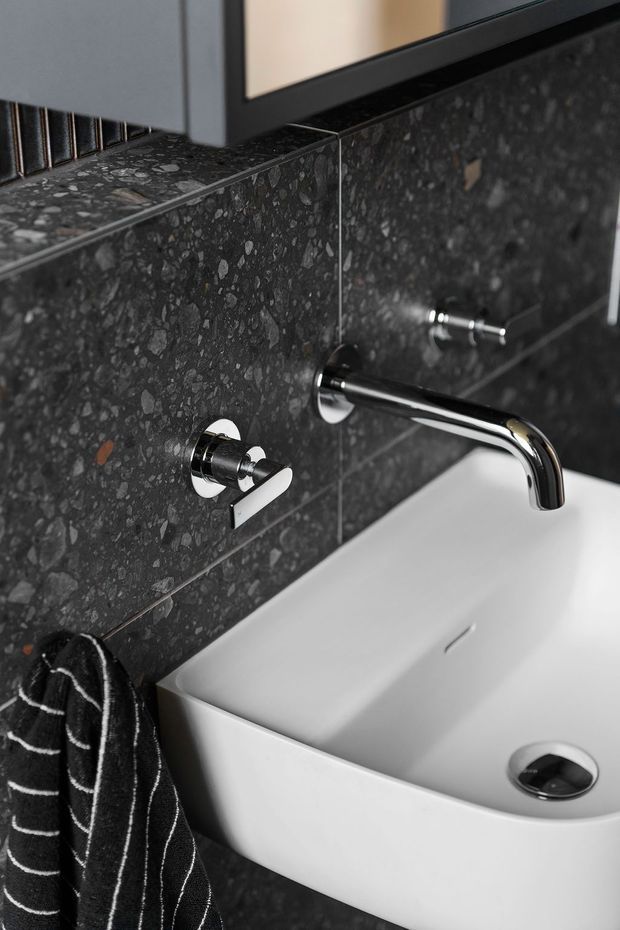 Close-up of a contemporary bathroom vanity featuring a white vessel basin, wall-mounted chrome tapware, and dark terrazzo-style stone wall tiles and benchtop. A black-and-white striped towel hangs at the side, with a partially visible mirror above.