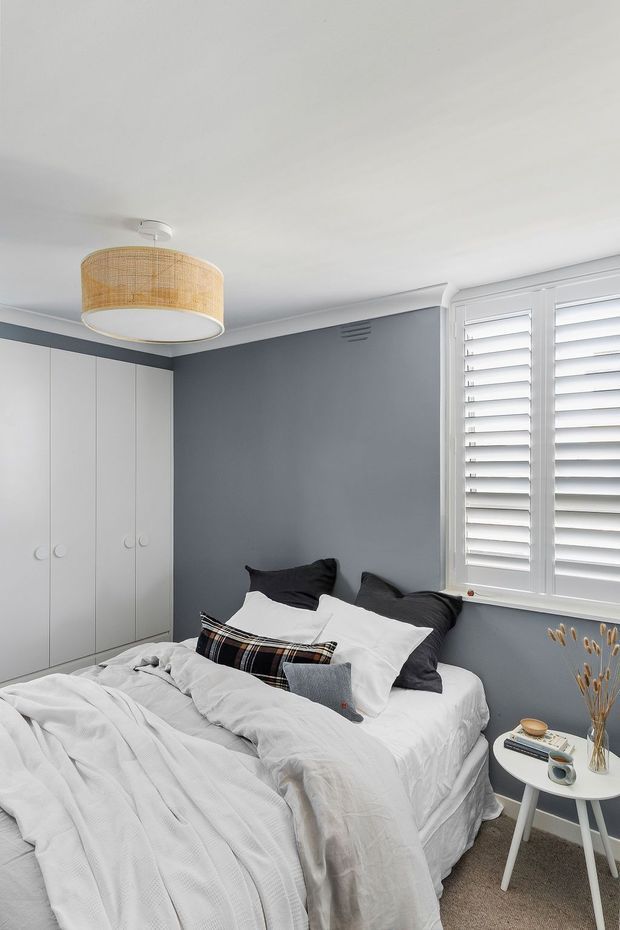 A compact, neatly styled bedroom with a white upholstered bed dressed in layered white bedding and dark charcoal cushions. Full-height white built-in wardrobes line the left wall, while a grey-blue feature wall and white plantation shutters frame the window. A woven rattan pendant light, small white bedside table, books, and dried decorative stems create a calm, contemporary interior.
