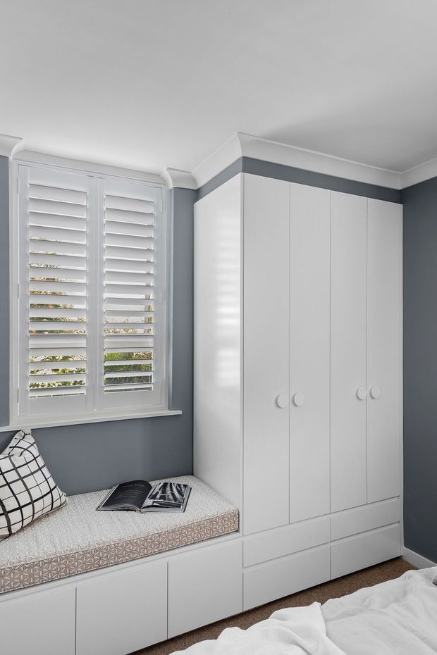 A compact, neatly designed bedroom with dark grey walls, white built-in wardrobe cabinetry, and a matching window seat beneath white plantation shutters. The built-in seat has a patterned upholstered cushion and a geometric accent pillow, while part of a white bed is visible in the foreground. The room has a clean, tailored and calm appearance.