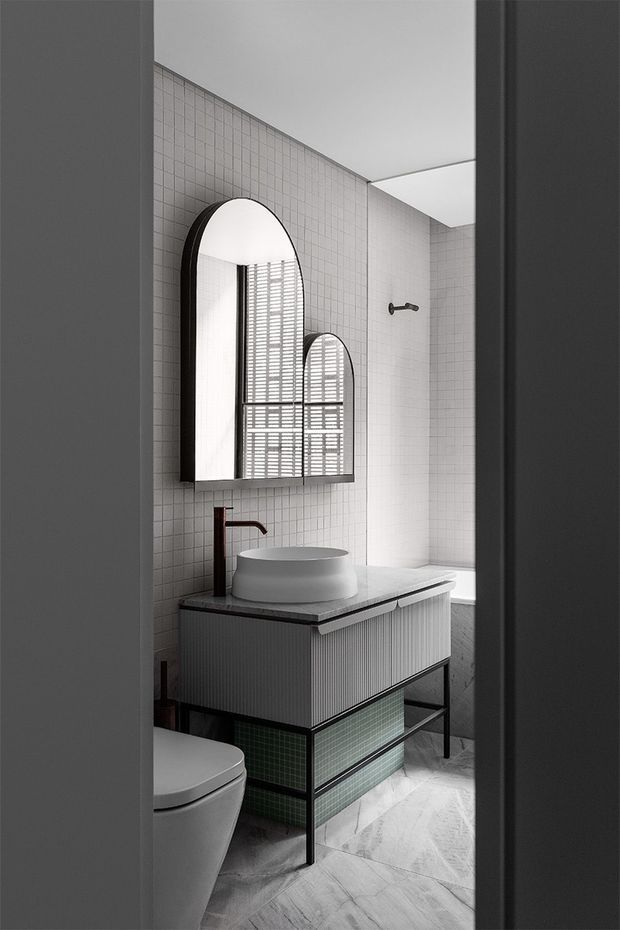 A compact contemporary bathroom viewed through a doorway, featuring white grid wall tiles, pale veined stone-look floor tiles, a ribbed floating vanity with a white vessel basin, dark metal legs and trim, green tiled lower storage, a dark faucet, and an arched mirrored cabinet. A toilet and shower area are also visible, with a bright skylight or ceiling opening above.