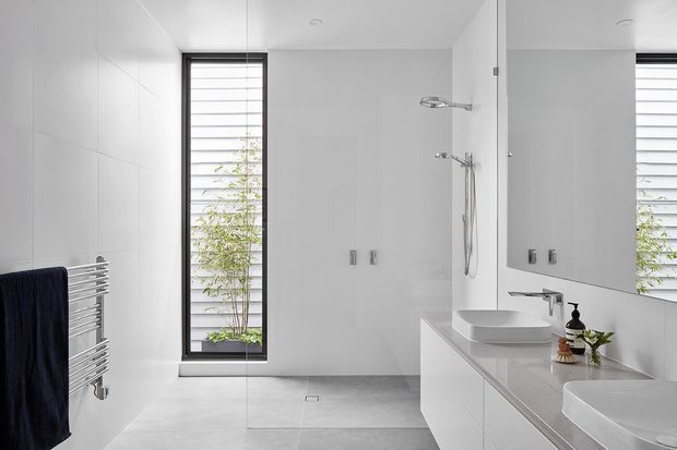 Bright contemporary bathroom with a walk-in glass shower, twin vessel basins on a long white vanity, large wall mirror, heated towel rail, pale tiled surfaces, and narrow black-framed louvre-style windows. The room has a clean, minimal, high-contrast monochrome appearance with natural greenery visible outside.
