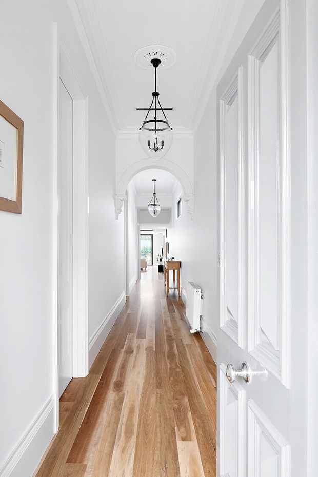 Bright, narrow residential hallway with white paneled walls, ornate ceiling cornices and decorative architraves, warm timber flooring, and a series of hanging glass pendant lights. A decorative arched opening leads toward a small console area and a bright glazed doorway at the far end.