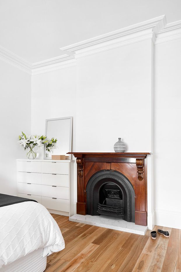 Bright, refined bedroom interior featuring ornate white wall mouldings and cornices, a restored dark timber mantel with a black decorative fireplace, white built-in or freestanding drawer storage, a leaning mirror, floral arrangement, and warm timber flooring. Part of a bed is visible in the foreground.