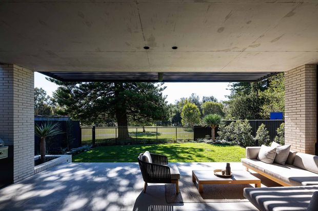 A covered outdoor living terrace opens onto a landscaped backyard. The space features a concrete ceiling and floor, pale brick side walls, dark horizontal roof louvers, a light upholstered sofa, timber coffee table, lounge chairs, and a view toward a fenced lawn and mature trees. The setting is bright and relaxed, with strong indoor-outdoor connection.