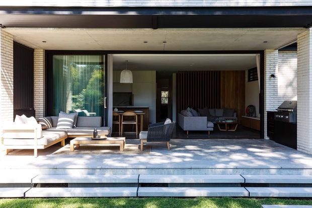 A covered outdoor terrace opens directly into a contemporary living room through wide sliding glazing. The terrace has pale stone or concrete paving, a low timber coffee table, light upholstered outdoor seating and a dark lounge chair. Inside, the living room features a grey sofa, dark timber vertical wall panelling, built-in joinery, and a dining or kitchen area beyond. White brick piers, dark horizontal roof framing, broad steps, and strong indoor-outdoor connection define the composition.