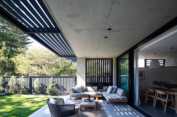 A contemporary covered outdoor terrace adjoining an open-plan dining area. The space features a concrete soffit, extensive dark-framed glazing, vertical slatted screens, a timber-toned outdoor lounge setting, and a retractable or louvred overhead shade structure. A landscaped garden is visible beyond the dark balustrade.