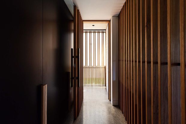 A narrow, refined residential hallway framed by dark timber doors and a full-height vertical timber batten screen. The corridor leads toward a bright glazed opening with additional vertical battens, creating strong linear perspective and filtered daylight. The floor appears to be a smooth light grey polished surface, while the walls and joinery are predominantly dark timber and neutral plaster.