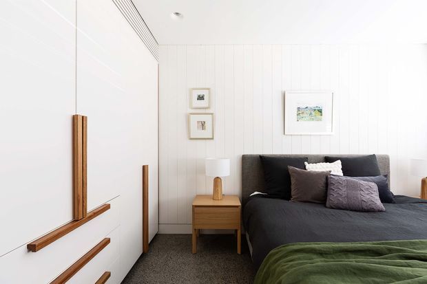 Bright, compact bedroom with a white vertical-panelled wall, large white built-in wardrobe cabinetry with warm timber handles and trim, and a dark upholstered bed layered with charcoal, mauve, and green bedding. A timber bedside table and simple lamp sit beside the bed, with small framed artwork adding detail. The room has a clean, contemporary coastal feel and soft natural light.