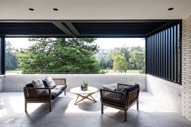 A bright, covered outdoor lounge terrace with contemporary architectural detailing. The space features a concrete floor and low parapet, dark louvred roof and vertical privacy screen, pale brickwork, and a relaxed arrangement of woven lounge chairs around a small round coffee table. Open views to landscaped greenery are framed through the terrace opening.