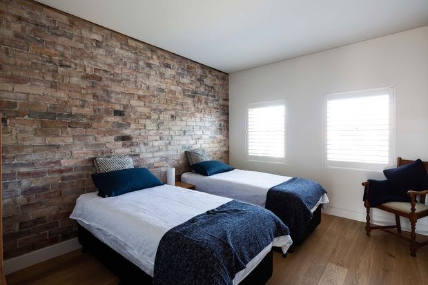 A bright bedroom with two single beds arranged parallel against a full-height exposed brick feature wall. The beds have white bedding with dark navy-blue throws and cushions, while a timber floor, white walls, plantation shutters, and a timber-framed armchair create a simple, warm, contemporary-rustic interior.