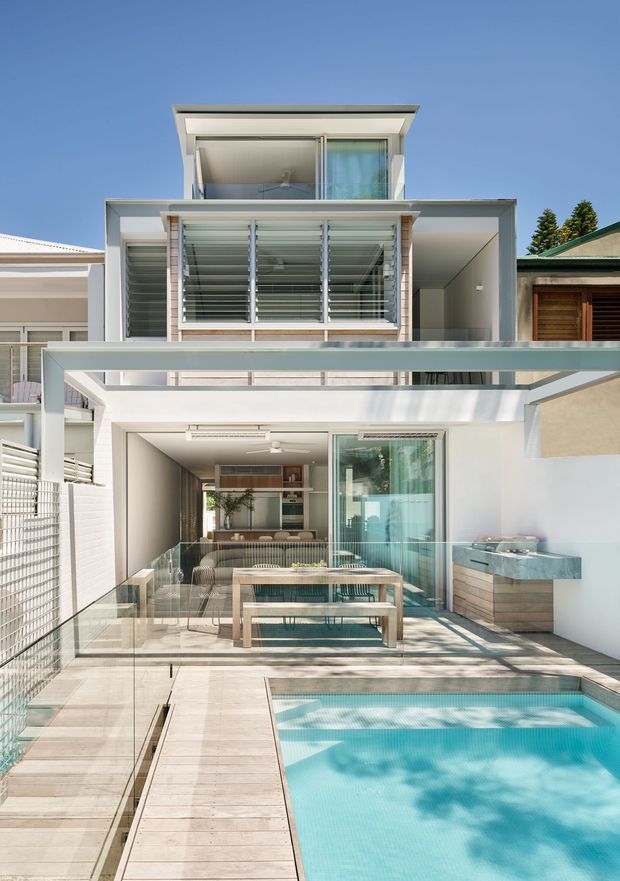 Exterior view of a contemporary multi-level residence with crisp white rendered walls, extensive glazing, horizontal louvres, and layered flat-roofed forms. A covered outdoor entertaining area contains a dining table and seating, with a built-in outdoor kitchen beside it. A glass balustrade overlooks a rectangular swimming pool edged in pale timber decking. The bright, sunlit composition shows a compact urban residential setting with strong indoor–outdoor connection.