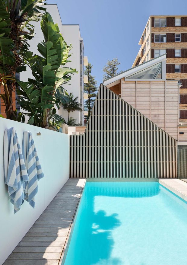 A bright urban residential courtyard with a narrow turquoise swimming pool, timber decking, pale rendered boundary walls, tropical foliage, and a distinctive slatted privacy screen with a steep triangular profile. A small glazed roof structure sits behind the screen, with multi-storey apartment buildings visible beyond. The scene has a clean contemporary coastal feel and strong geometric lines.