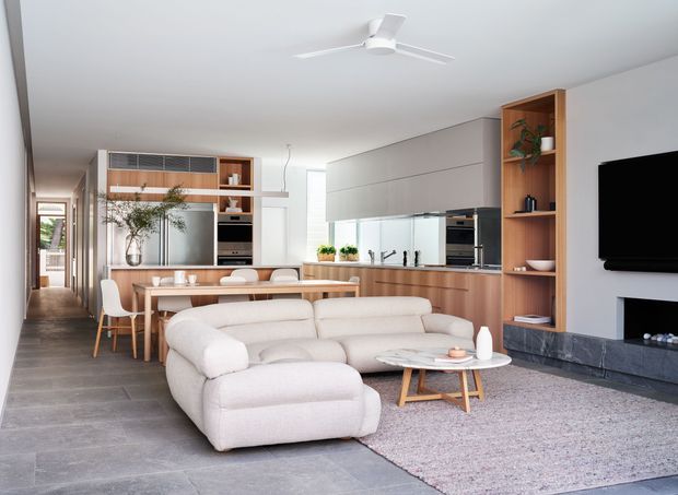 Open-plan contemporary living, dining and kitchen area with a light neutral palette. The room features a large modular cream sofa, timber dining table and chairs, a linear kitchen with pale cabinetry and timber accents, integrated appliances, open shelving, grey tiled flooring, a textured rug, and a built-in TV/fireplace wall. A hallway extends behind the dining area, and a ceiling fan hangs from the smooth white ceiling.