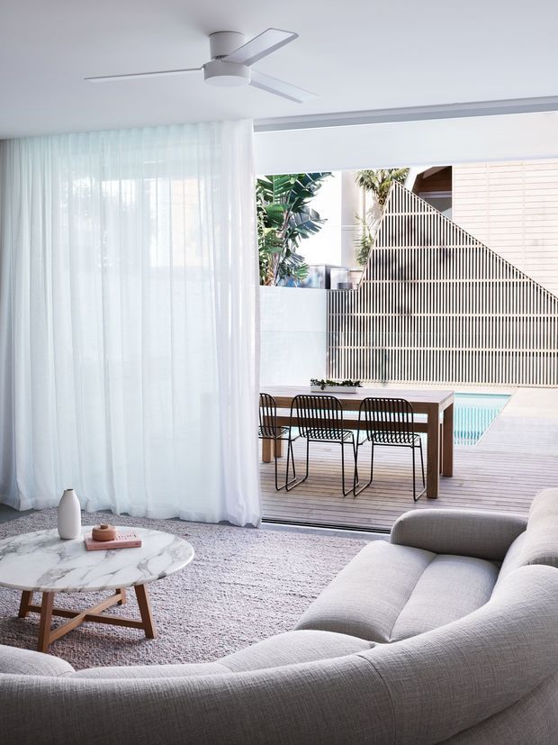 Bright contemporary living room opening onto a timber deck and pool area. The room features a large curved light-grey sectional sofa, pale textured rug, round marble coffee table with timber legs, sheer white curtains, and a minimalist white ceiling fan. Outside, a timber dining table with black wire chairs sits beside a pool, framed by a distinctive slatted privacy screen and tropical planting.